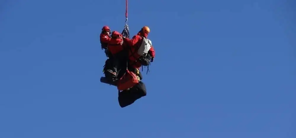 A rescuer suspended on a long line beneath a helicopter at dusk